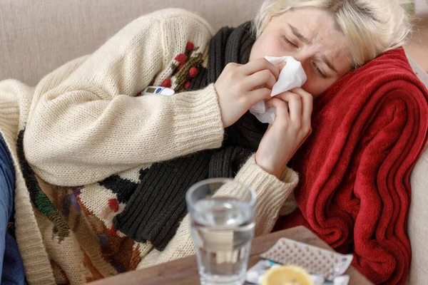 Дівчина в светрі чхає, сидячи на дивані. Застуда і грип. Пацієнт застудився, почувався хворим і чхав у паперовій серветці. Здорова дівчина витерла їй ніс . — стокове фото