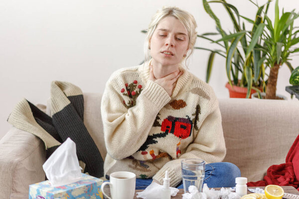 The girl in the sweater is sick, sitting on the sofa. Colds and flu. The patient caught a cold, feeling sick and sore throat. Unhealthy girl with a sore throat.