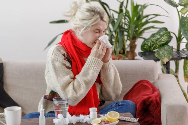 Chica en un suéter estornuda mientras está sentado en un sofá. El paciente se resfrió, sintiéndose enfermo y estornudando en una servilleta de papel. Una chica malsana se limpió la nariz . —  Fotos de Stock