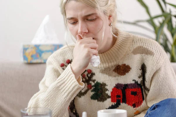 The girl in the sweater coughs, sitting on the couch. Colds and flu. The patient caught a cold, feeling sick and coughing in a napkin. An unhealthy girl coughs.