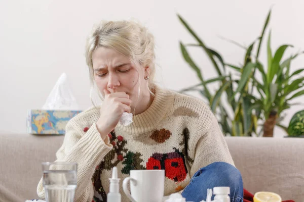 A lány a pulcsiban köhög, a kanapén ül. Megfázás és influenza. A beteg megfázott, rosszul lett és köhög egy szalvétában. Egy egészségtelen lány köhög.. — Stock Fotó