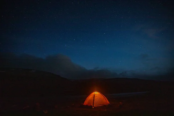 Dramatik akşam gökyüzünün altındaki dağlarda parlayan turuncu çadır. Kırmızı günbatımı ve arka planda dağlar. Yaz manzarası.