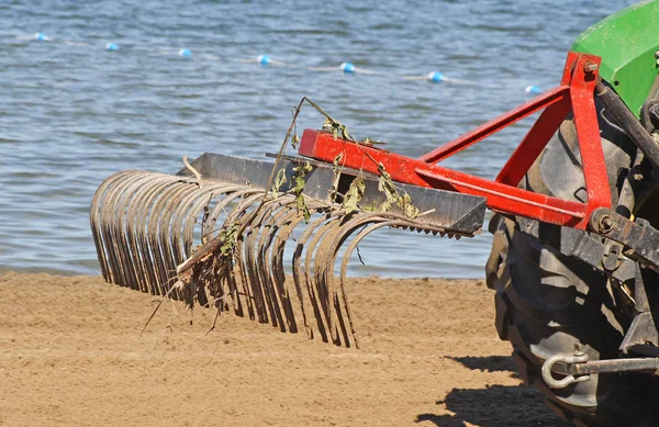 大きな金属熊手が取り付けられたトラクターは 早朝に公共のビーチでふるみ レーキング 破片を取り除きます ロイヤリティフリーのストック画像