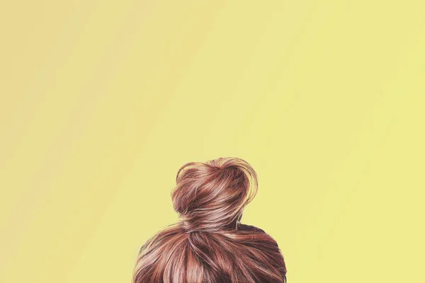 A view of the back of a woman's head. Hair wrapped in a bun on a light yellow pastel background. Content completion concept.