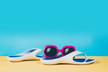 Parmak arası terlik, sandalet, güneş gözlüğü ve deniz kabuğu, yaz seti mavi. Yaz önümüzdeki kavramdır. En az style, minimalist fotoğraf. Pastel renkler sarı, pembe ve mavi arka plan.