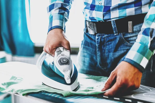 Man at home ironing clothes. Father performs home duties, irons the child's clothes. Man in role of a woman at home. Man ironing shirt on ironing board with steaming blue iron.