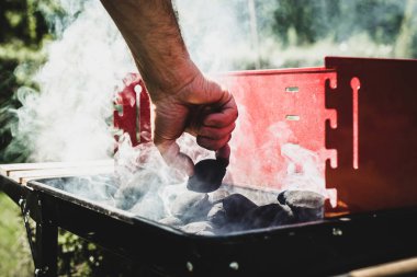 Izgara aydınlatma. Adam mangal yakmak. Elini kömür ızgara için ekliyor. Grill'de aile ile birlikte vakit. Sosyal toplantılar, arkadaşlar. Odun ve kömür ile kırmızı kiremit.