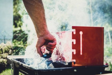 Izgara aydınlatma. Adam mangal yakmak. Elini kömür ızgara için ekliyor. Grill'de aile ile birlikte vakit. Sosyal toplantılar, arkadaşlar. Odun ve kömür ile kırmızı kiremit.