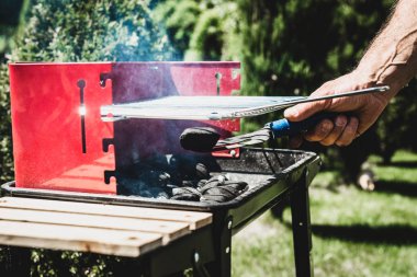 Izgara aydınlatma. Adam mangal yakmak. Elini kömür ızgara için ekliyor. Grill'de aile ile birlikte vakit. Sosyal toplantılar, arkadaşlar. Odun ve kömür ile kırmızı kiremit.