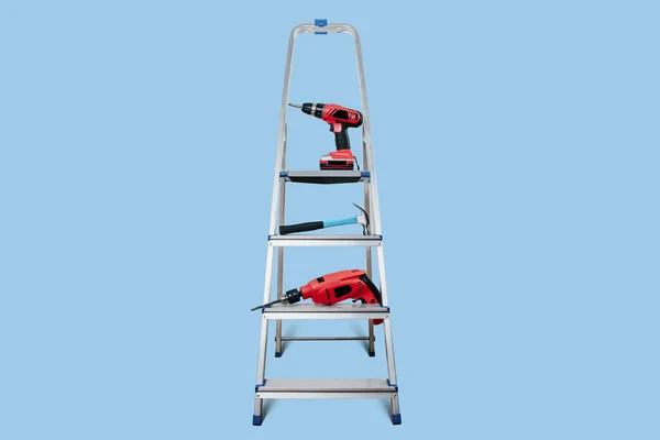 A view of construction tools. A cordless screwdriver, a drill and a hammer are arranged on the ladder. Tools necessary at home on a light blue pastel background.
