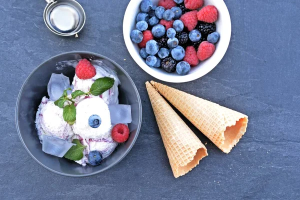 A dark bowl full of wild berry ice cream balls with mint leaves and ice cream waffles next to it on a dark kitchen surface - concept with refreshing fruit ice for the summer