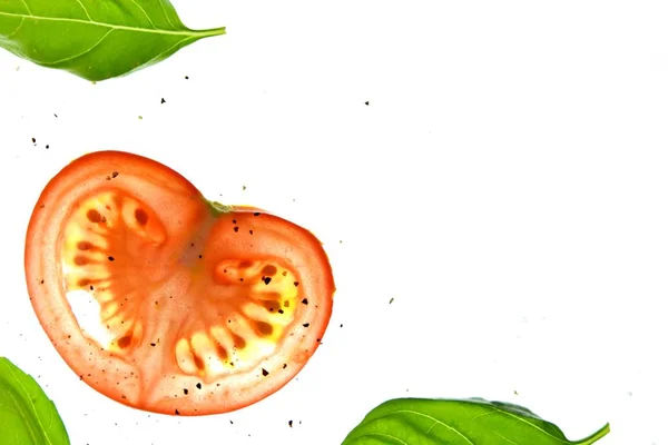 Sliced tomato pieces lay with basil leaves on a white bright background illuminated from above and sprinkled with spices as salt and pepper