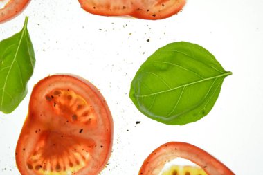 Dilimlenmiş domates parçaları yukarıdan aydınlatılmış beyaz parlak bir arka plan üzerinde fesleğen yaprakları ile yatıyordu ve tuz ve biber gibi baharat serpilir