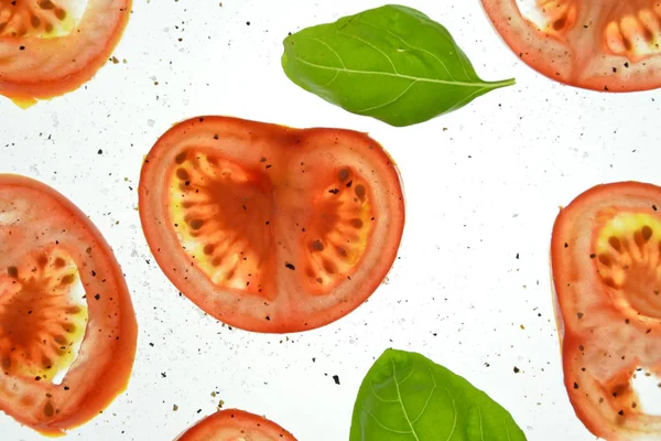 Sliced tomato pieces lay with basil leaves on a white bright background illuminated from above and sprinkled with spices as salt and pepper