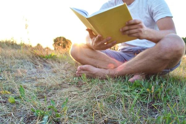 Yakın çekim - güneş bir ormanın yakınında arka planda batarken ayakkabı ve şort olmadan kırsal bir kitap okuma adam - okuma sevgisi için kavram 