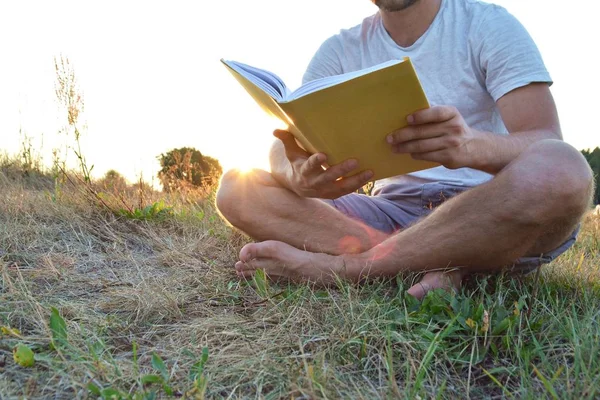 Yakın çekim - güneş bir ormanın yakınında arka planda batarken ayakkabı ve şort olmadan kırsal bir kitap okuma adam - okuma sevgisi için kavram 
