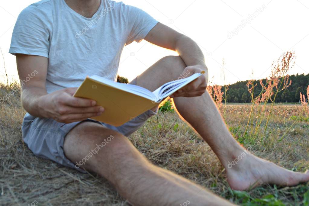 Close-up - man reading a book in the countryside without shoes and shorts while the sun is setting in the background near a forest - concept for the love of reading 