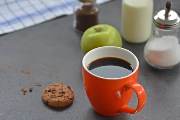 Coffee in an orange cup on a table with apples, milk, sugar and a biscuit