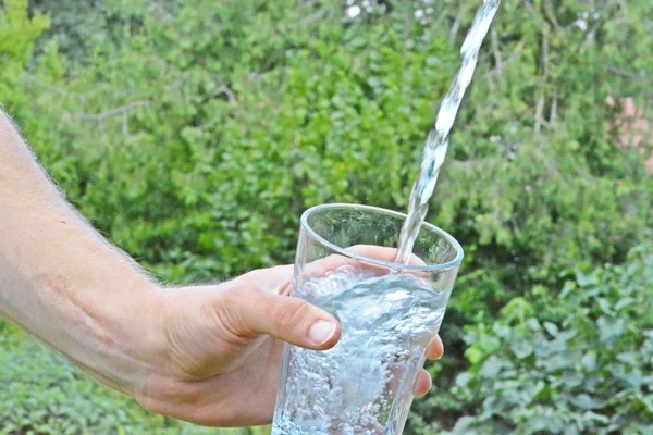 新鲜干净的水在绿色的夏日背景前的玻璃杯中流动 由一个人的手拿着 — 图库照片
