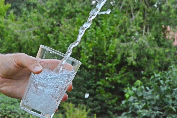 Fresh and clean water flows in a glass in front of a green summery background and held by a man\'s hand