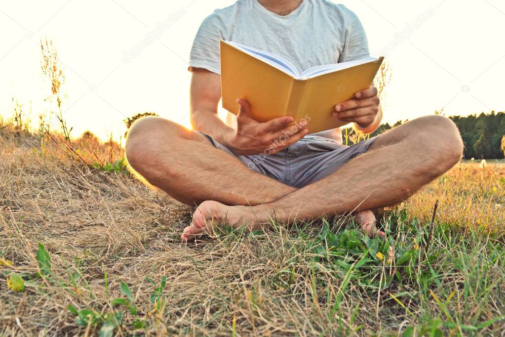 Close-up - man reading a book in the countryside without shoes and shorts while the sun is setting in the background near a forest - concept for the love of reading 