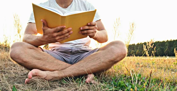 Yakın çekim - güneş bir ormanın yakınında arka planda batarken ayakkabı ve şort olmadan kırsal bir kitap okuma adam - okuma sevgisi için kavram 