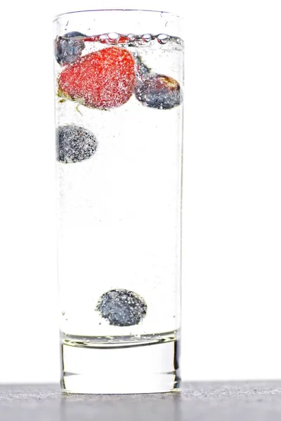 スパークリングウォーターでいっぱいのグラスにブルーベリーとイチゴを落とす 隔離されたガラスの上に閉じる — ストック写真