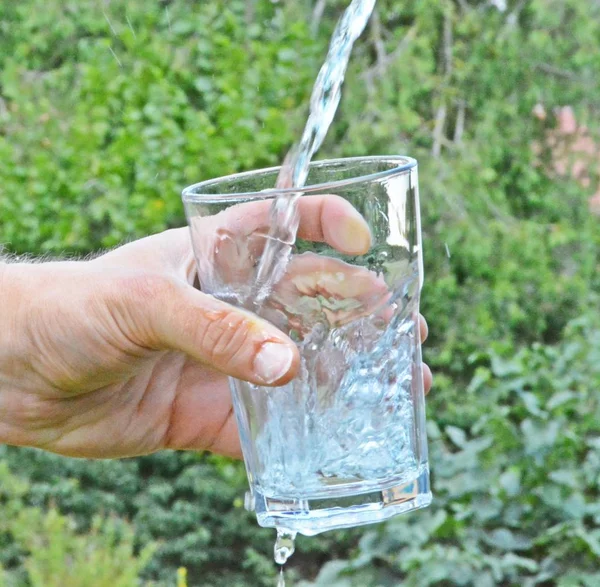 Fresh and clean water flows in a glass in front of a green summery background and held by a man\'s hand