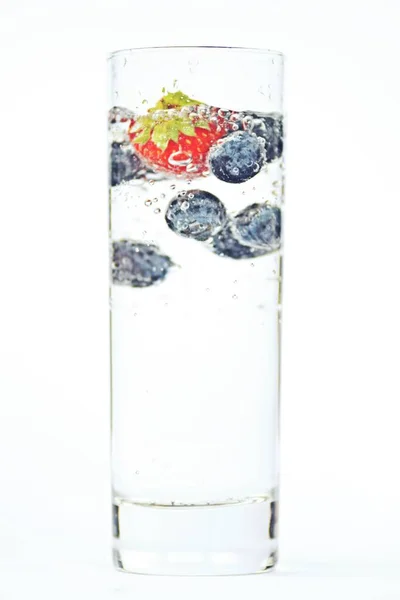 スパークリングウォーターでいっぱいのグラスにブルーベリーとイチゴを落とす 隔離されたガラスの上に閉じる — ストック写真