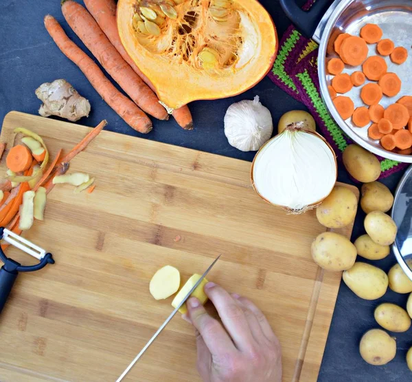 Cutting vegetables for carrots, potatoes, onions, garlic and a pumpkin for pumpkin soup on a wooden board and pouring into the pot - closeup on the hands from above