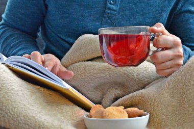 Genç bir adam bir kanepe üzerinde oturuyor ve el ve bisküvi onun yanında stand bir cam bardak bir meyve çayı tutarken bir kitap okuma-kitap ve cam fincan çay dolu-kavramı ısınmak için