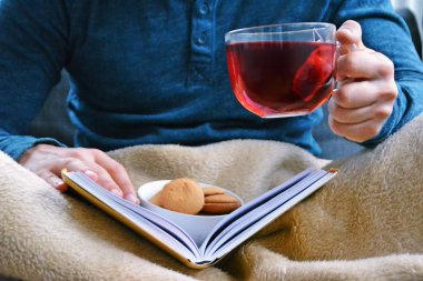 Genç bir adam bir kanepe üzerinde oturuyor ve el ve bisküvi onun yanında stand bir cam bardak bir meyve çayı tutarken bir kitap okuma-kitap ve cam fincan çay dolu-kavramı ısınmak için