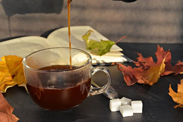 Pour coffee in a autumn setting into a glass cup - a glass cup is standig beside dry and colorful autumn leaves and a book in the background and someone is pouring coffee into it