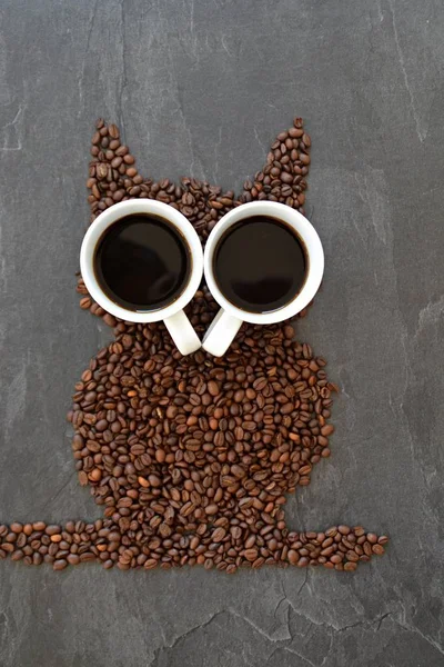 Coffee beans on a black background form the silhouette of an owl and two cups of coffee form their eyes - concept with coffee and night owls