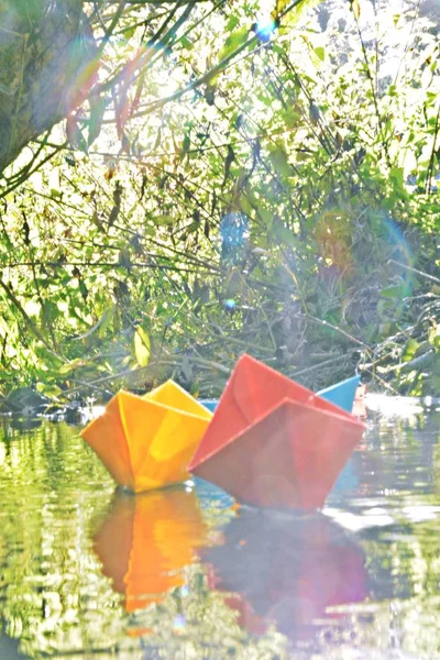 Different colored paper ships swim in a small brook on the surface of which reflects the late autumn sun - Slightly blurred paper ships in bright colors on a lake in late autumn