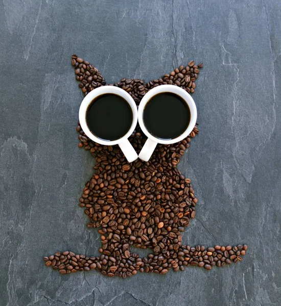 Coffee beans on a black background form the silhouette of an owl and two cups of coffee form their eyes - concept with coffee and night owls