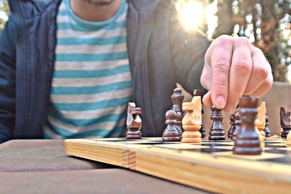 A man in his mid-twenties sits in an autumnal park and plays chess - focusing on the game pieces on the board, the person can not be seen and only the hands and torso are visible