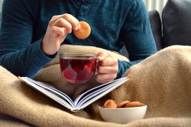 Genç bir adam bir kanepe üzerinde oturuyor ve el ve bisküvi onun yanında stand bir cam bardak bir meyve çayı tutarken bir kitap okuma-kitap ve cam fincan çay dolu-kavramı ısınmak için