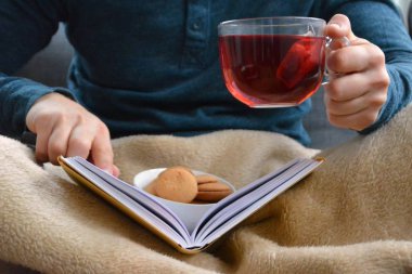 Genç bir adam bir kanepe üzerinde oturuyor ve el ve bisküvi onun yanında stand bir cam bardak bir meyve çayı tutarken bir kitap okuma-kitap ve cam fincan çay dolu-kavramı ısınmak için