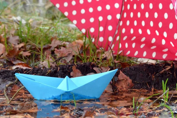 A self-folded paper boat floats in a puddle in nature and is greeted by a pink umbrella with white dots - autumn scenario with paper boat in nature