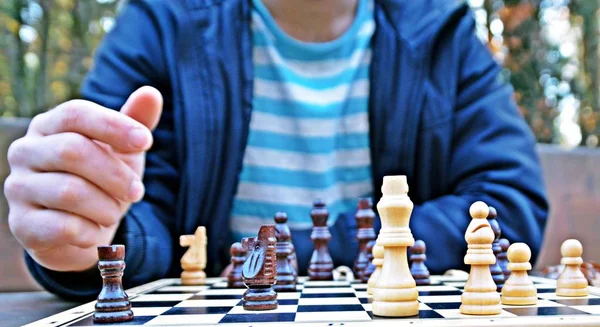 A man in his mid-twenties sits in an autumnal park and plays chess - focusing on the game pieces on the board, the person can not be seen and only the hands and torso are visible