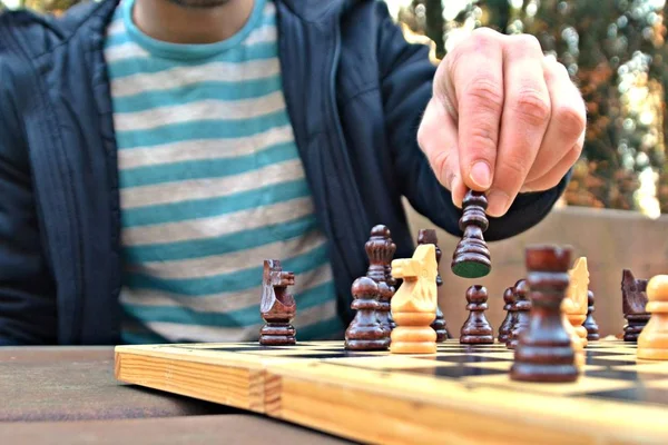 A man in his mid-twenties sits in an autumnal park and plays chess - focusing on the game pieces on the board, the person can not be seen and only the hands and torso are visible