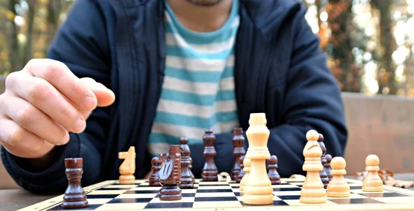 A man in his mid-twenties sits in an autumnal park and plays chess - focusing on the game pieces on the board, the person can not be seen and only the hands and torso are visible