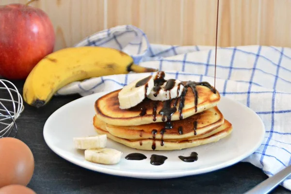 Pancakes with bananas and chocolate sauce on a black table with a wooden background, together with fruit, eggs and a blue white checked kitchen towel