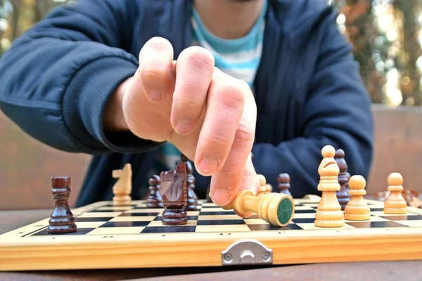 A man in his mid-twenties sits in an autumnal park and plays chess - focusing on the game pieces on the board, the person can not be seen and only the hands and torso are visible