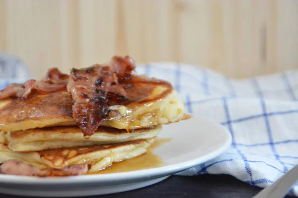 Pancakes with bananas and chocolate sauce on a black table with a wooden background, together with fruit, eggs and a blue white checked kitchen towel