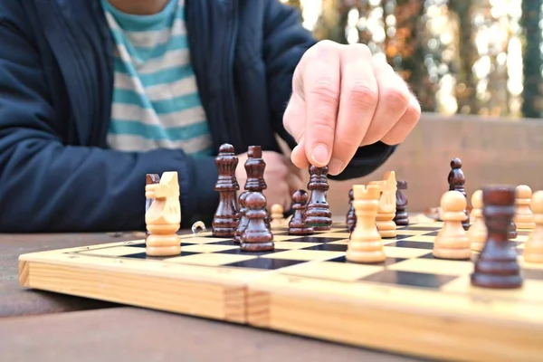 A man in his mid-twenties sits in an autumnal park and plays chess - focusing on the game pieces on the board, the person can not be seen and only the hands and torso are visible