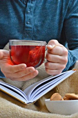 Genç bir adam bir kanepe üzerinde oturuyor ve el ve bisküvi onun yanında stand bir cam bardak bir meyve çayı tutarken bir kitap okuma-kitap ve cam fincan çay dolu-kavramı ısınmak için