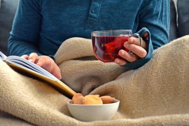 Genç bir adam bir kanepe üzerinde oturuyor ve el ve bisküvi onun yanında stand bir cam bardak bir meyve çayı tutarken bir kitap okuma-kitap ve cam fincan çay dolu-kavramı ısınmak için