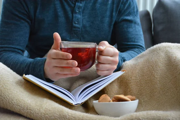 Genç bir adam bir kanepe üzerinde oturuyor ve el ve bisküvi onun yanında stand bir cam bardak bir meyve çayı tutarken bir kitap okuma-kitap ve cam fincan çay dolu-kavramı ısınmak için
